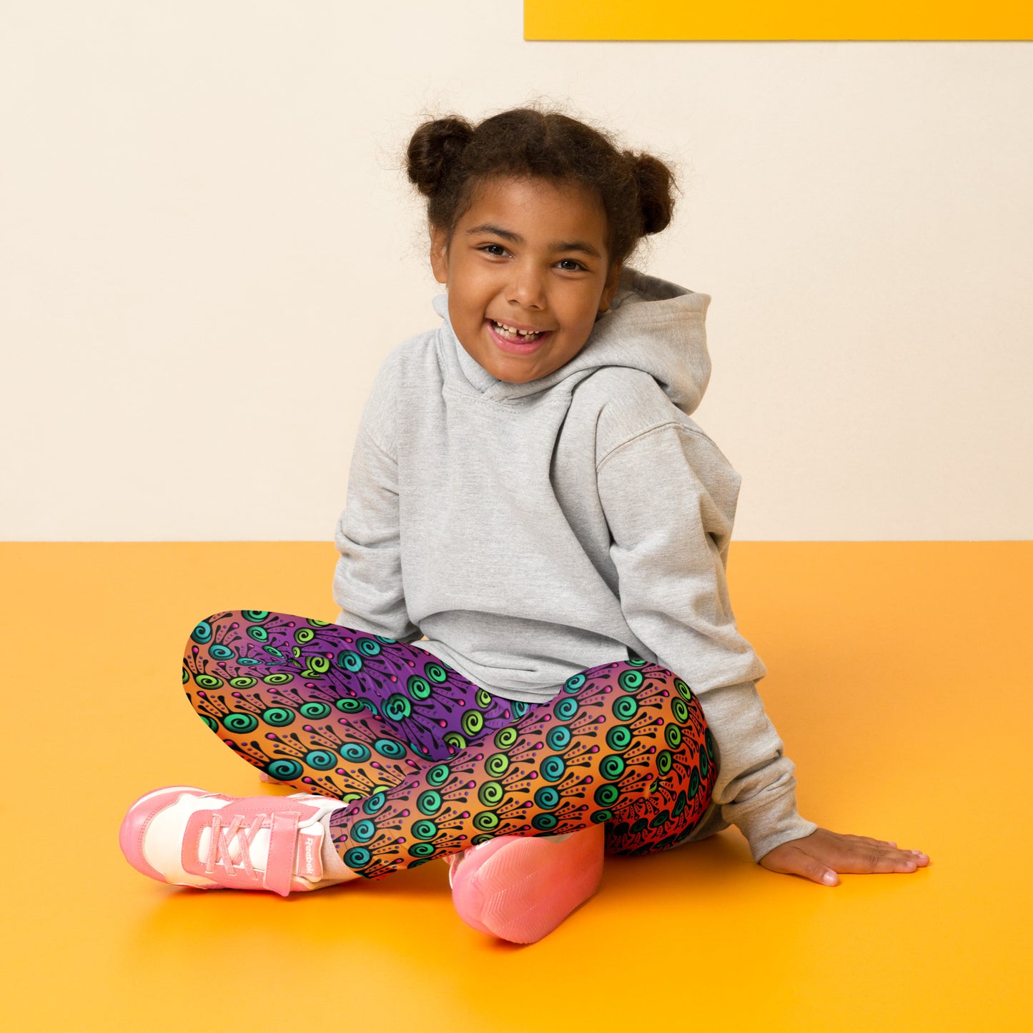 A young girl sits crossed legged wearing kids size fire tribal frog funky festival leggings and a light grey hoodie. She is sitting on a yellow floor against a cream wall. The fire tribal frog pattern  is an abstract pattern with a blended orange, pink and purple background.