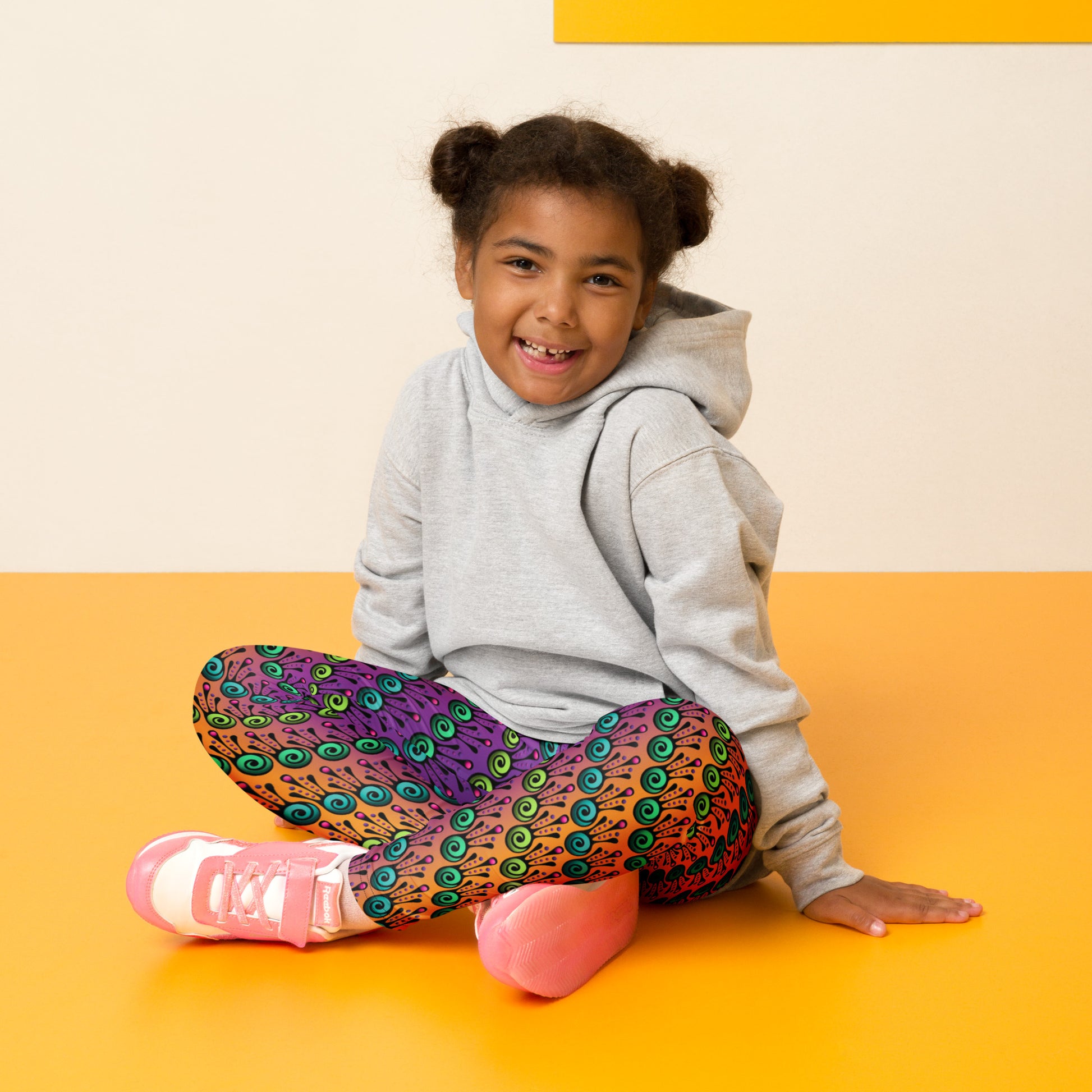 A young girl sits crossed legged wearing kids size fire tribal frog funky festival leggings and a light grey hoodie. She is sitting on a yellow floor against a cream wall. The fire tribal frog pattern  is an abstract pattern with a blended orange, pink and purple background.