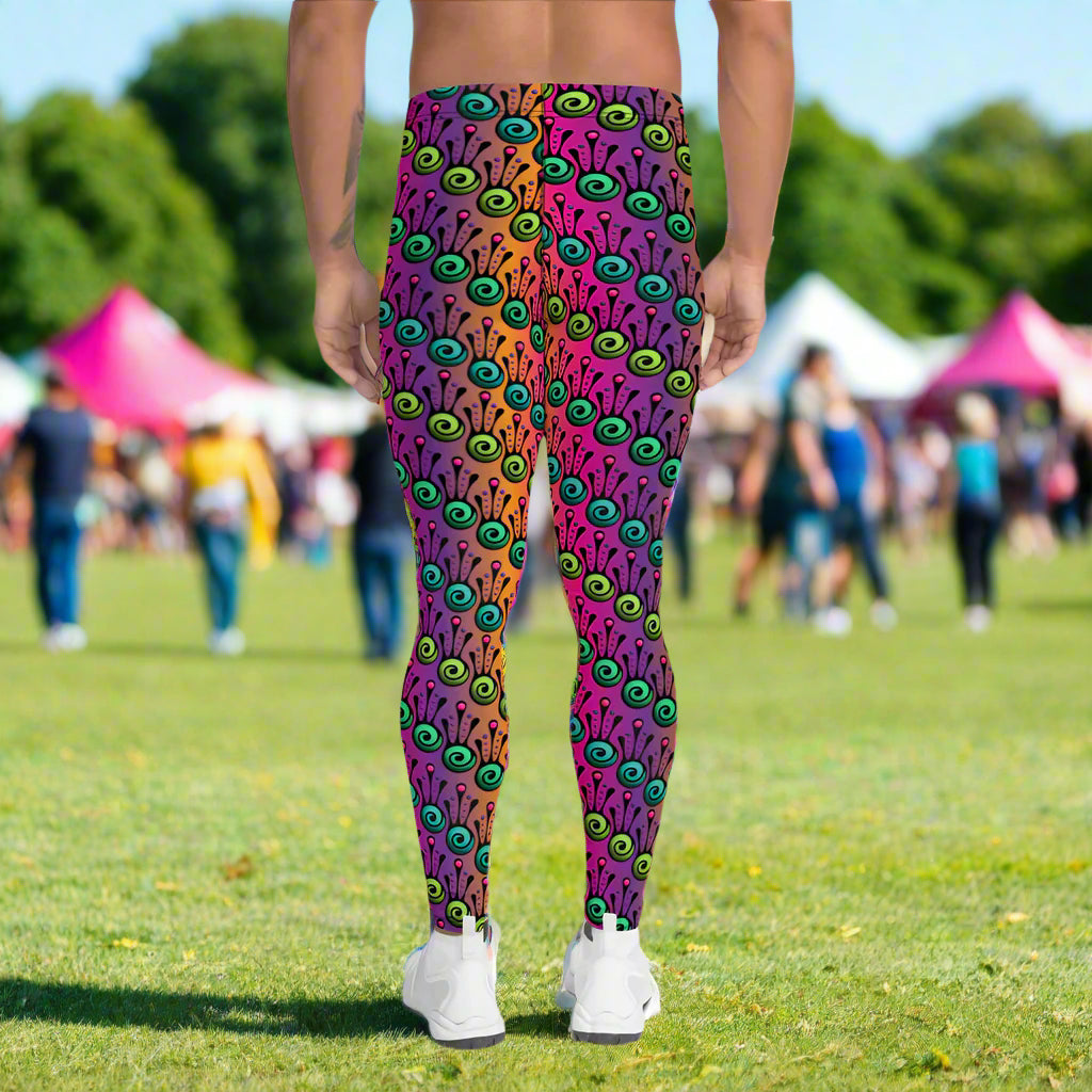 Man from the waist down wearing men's fire tribal frog funky festival yoga leggings standing facing away with his hands by his sides. He is wearing white trainers and is standing in a grassy festival field with other festival goers and market gazebos in the background. It is a sunny day. The fire tribal frog pattern  is a funky abstract pattern with a blended pink, purple and orange background.