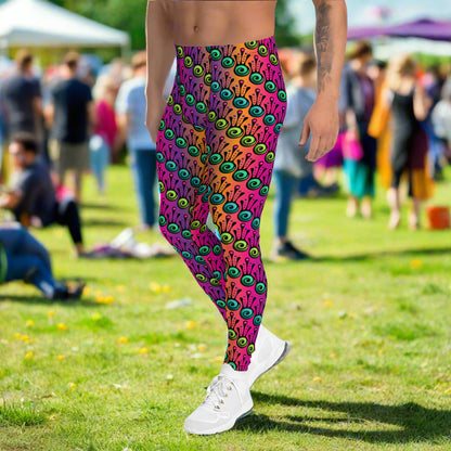 Man from the waist down wearing men's fire tribal frog funky festival yoga leggings standing facing to the left with his hands by his sides. He is wearing white trainers and is standing in a grassy festival field with other festival goers mingling in the background. It is a sunny day. The fire tribal frog pattern  is a funky abstract pattern with a blended pink, purple and orange background.