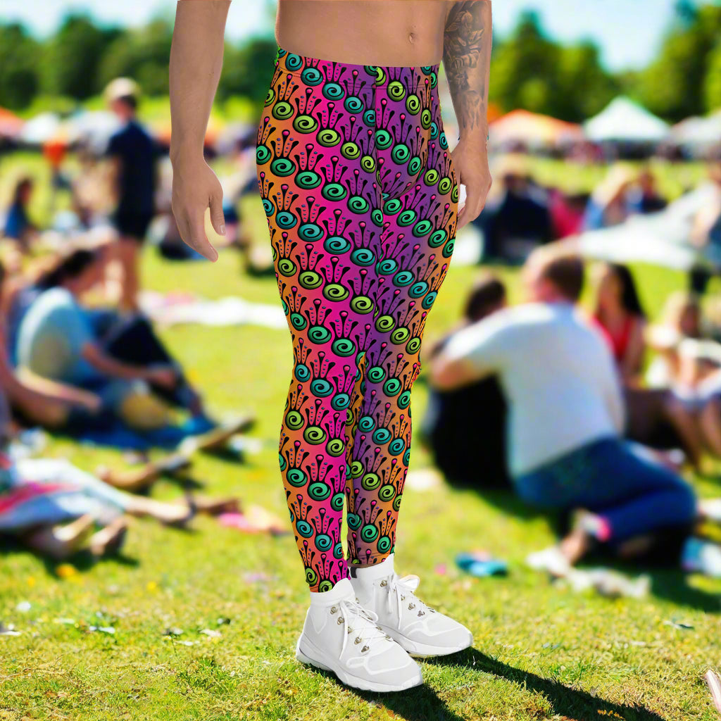 Man from the waist down wearing men's fire tribal frog funky festival yoga leggings standing facing to the right with his hands by his sides. He is wearing white trainers and is standing in a grassy festival field with other festival goers sitting around on the grass in the background. It is a sunny day. The fire tribal frog pattern  is a funky abstract pattern with a blended pink, purple and orange background.