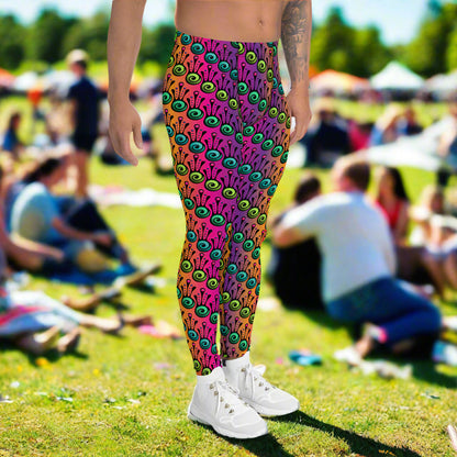 Man from the waist down wearing men's fire tribal frog funky festival yoga leggings standing facing to the right with his hands by his sides. He is wearing white trainers and is standing in a grassy festival field with other festival goers sitting around on the grass in the background. It is a sunny day. The fire tribal frog pattern  is a funky abstract pattern with a blended pink, purple and orange background.