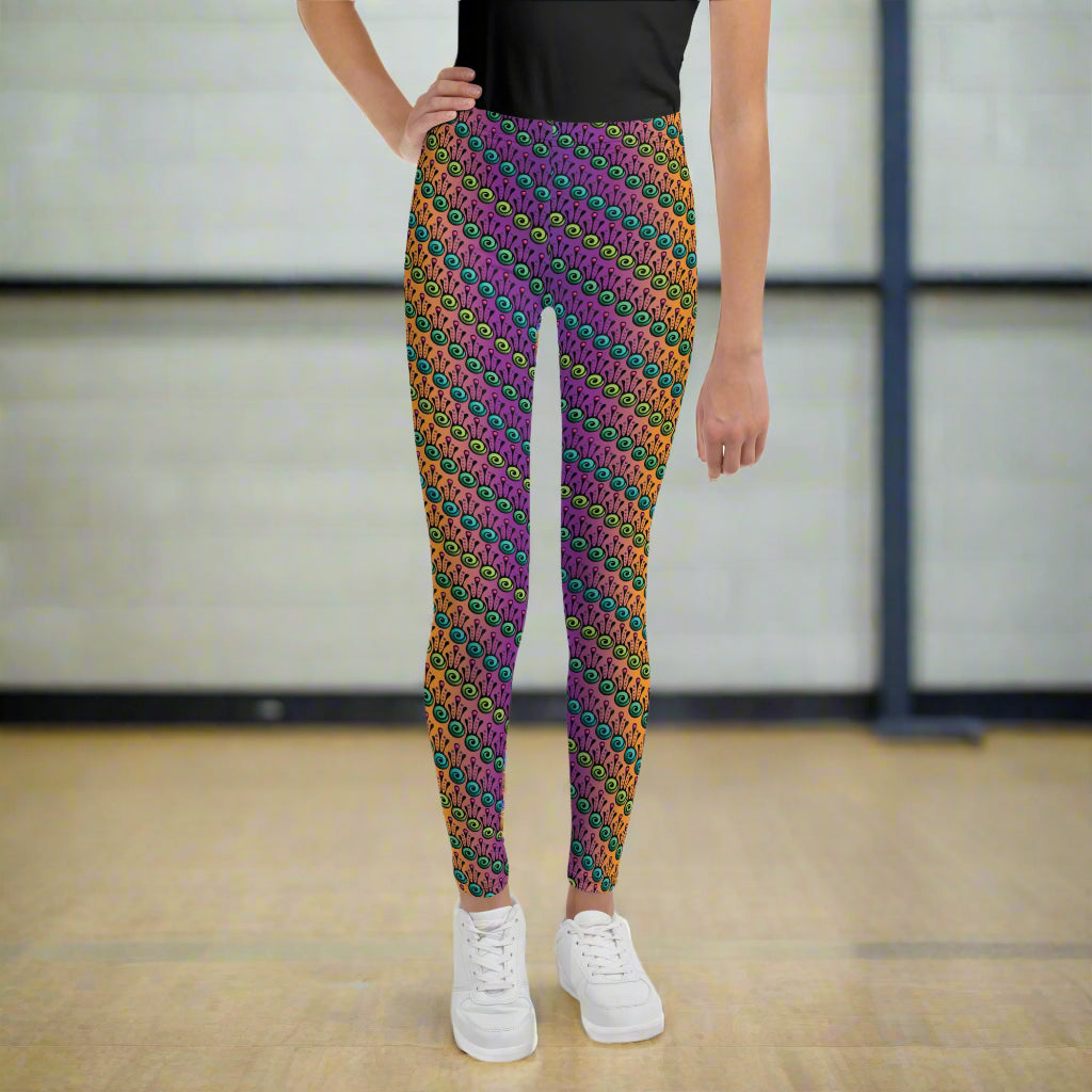 A youth pictured from the waist down wearing youth size fire tribal frog funky festival leggings standing facing forward with one hand on their hip. They are wearing white trainers and standing on a wooden gymnasium floor with a cream wall. The fire tribal frog pattern  is an abstract pattern with a blended orange, pink and purple background.