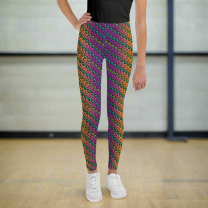 A youth pictured from the waist down wearing youth size fire tribal frog funky festival leggings standing facing forward with one hand on their hip. They are wearing white trainers and standing on a wooden gymnasium floor with a cream wall. The fire tribal frog pattern  is an abstract pattern with a blended orange, pink and purple background.