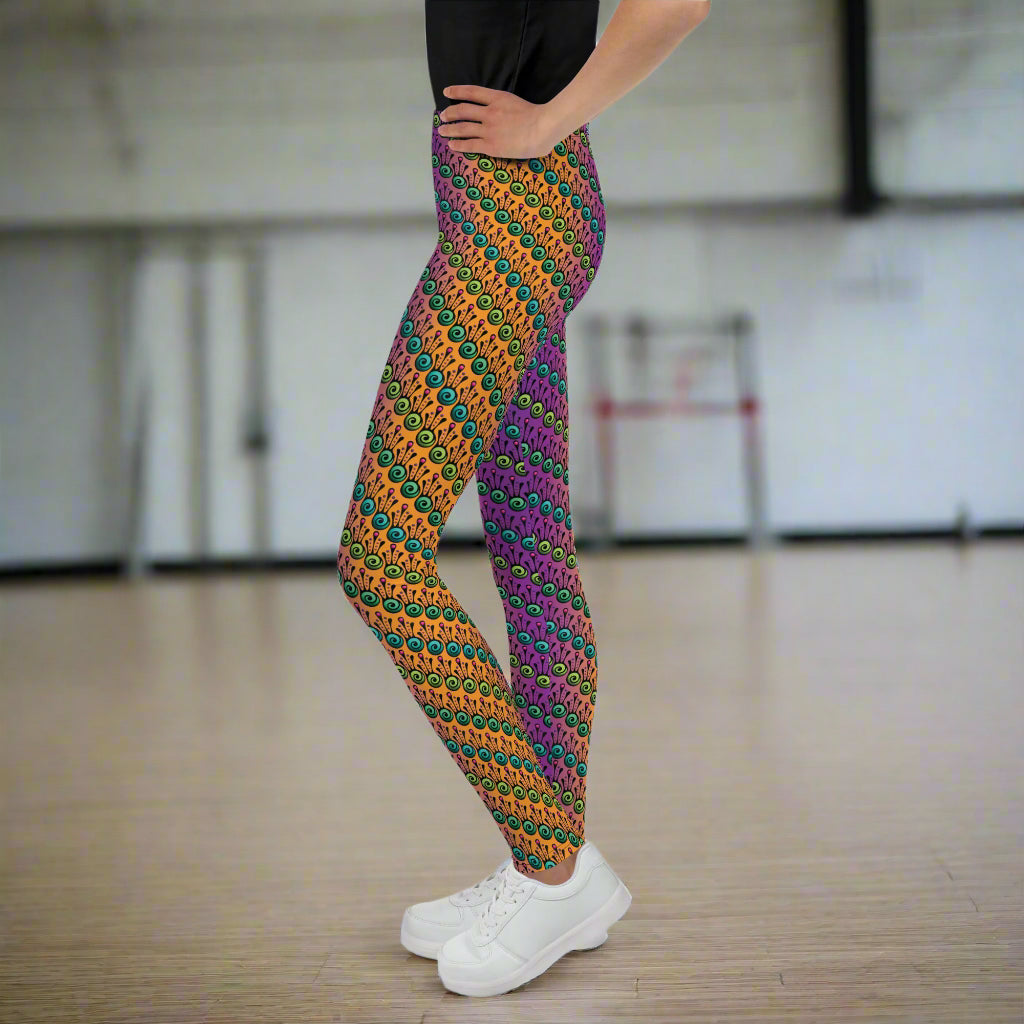 A youth pictured from the waist down wearing youth size fire tribal frog funky festival leggings standing facing to the left with both hands on their hips. They are wearing white trainers and standing on a wooden gymnasium floor with a cream wall. The fire tribal frog pattern  is an abstract pattern with a blended orange, pink and purple background.