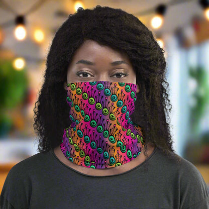 Woman with dark afro hair facing to the front wearing a dark grey long sleeve top and a vibrant tribal frog neck gaiter around her neck, covering her nose and mouth. In the background are some fairy lights and plants. The fire tribal frog design is an abstract pattern with a blended purple, pink and orange background. The abstract detail is coloured in different shades of green.