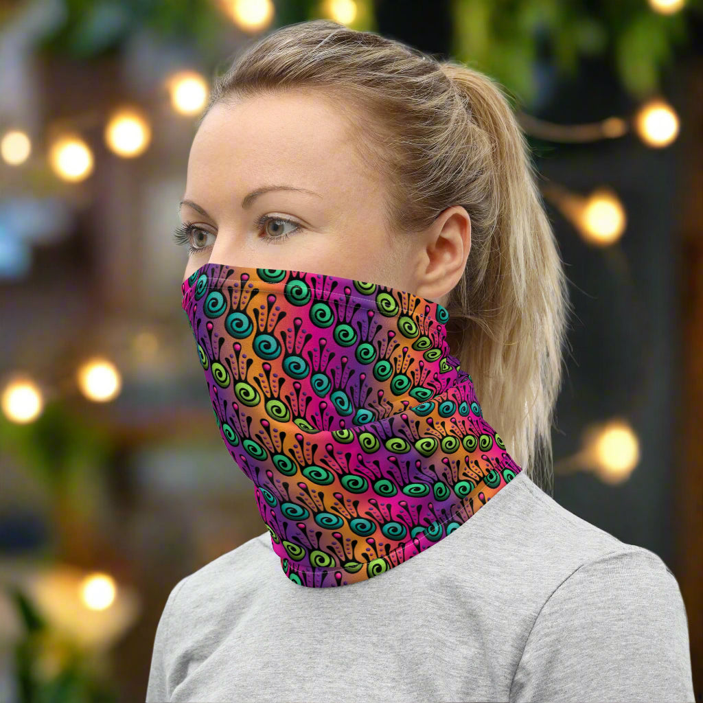 Woman with blonde hair tied up in a pony tail facing to the left wearing a grey long sleeve top and a vibrant tribal frog neck gaiter around her neck, covering her nose and mouth. In the background are some fairy lights and plants. The fire tribal frog design is an abstract pattern with a blended purple, pink and orange background. The abstract detail is coloured in different shades of green.