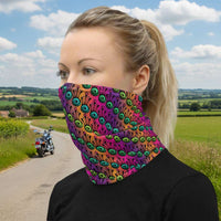 Woman with blonde hair tied up in a pony tail facing to the left wearing a grey long sleeve top and a vibrant tribal frog neck gaiter around her neck, covering her nose and mouth. In the background are some fairy lights and plants. The fire tribal frog design is an abstract pattern with a blended purple, pink and orange background. The abstract detail is coloured in different shades of green.