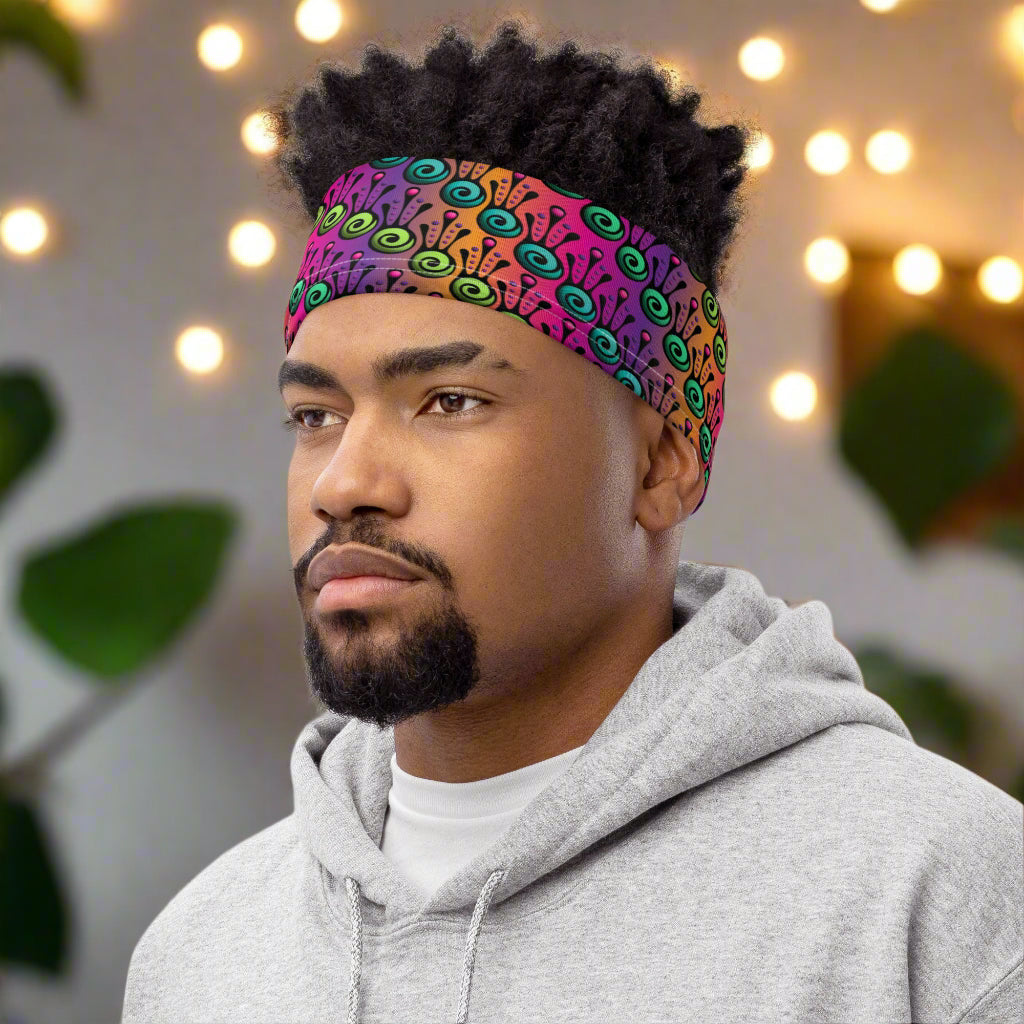 Man with short, dark, afro hair is  facing to the left wearing a grey hoodie and a vibrant tribal frog neck gaiter around his head, as a head band. In the background are some fairy lights and plants. The fire tribal frog design is an abstract pattern with a blended purple, pink and orange background. The abstract detail is coloured in different shades of green.