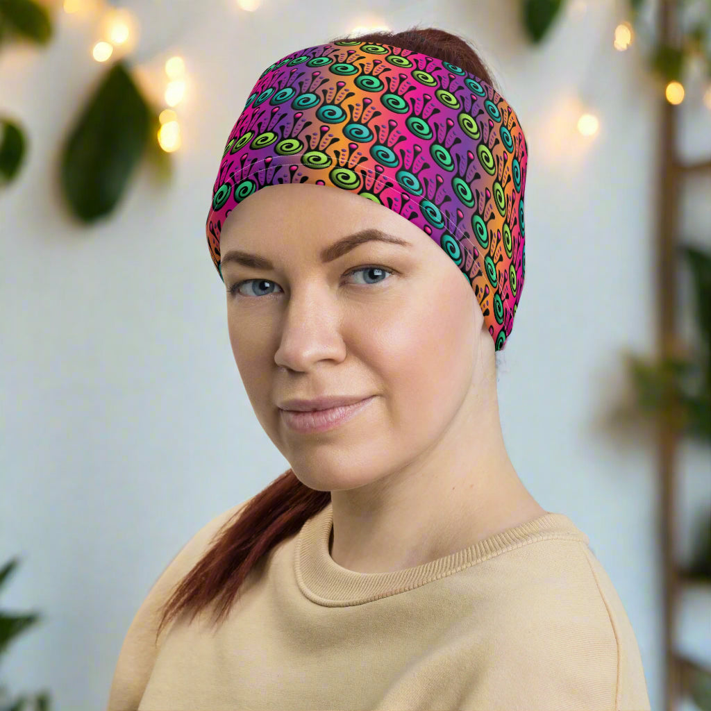Woman with red hair tied up in a pony tail facing to the left wearing a beige long sleeve top and a vibrant tribal frog neck gaiter around her head as a wide head band. In the background are some fairy lights and plants. The fire tribal frog design is an abstract pattern with a blended purple, pink and orange background. The abstract detail is coloured in different shades of green.