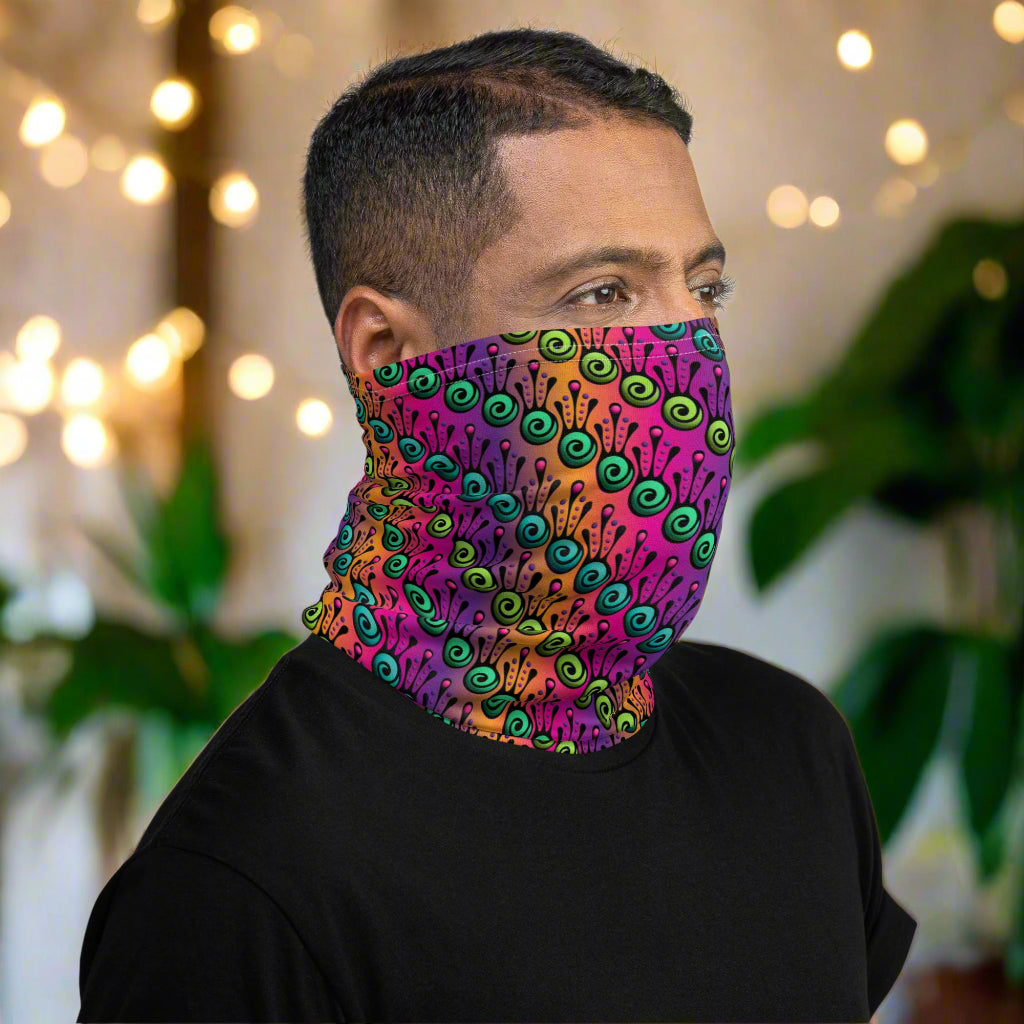 Man with short, dark hair facing to the right wearing a black long sleeve top and a vibrant tribal frog neck gaiter around his neck, covering his nose and mouth. In the background are some fairy lights and plants. The fire tribal frog design is an abstract pattern with a blended purple, pink and orange background. The abstract detail is coloured in different shades of green.