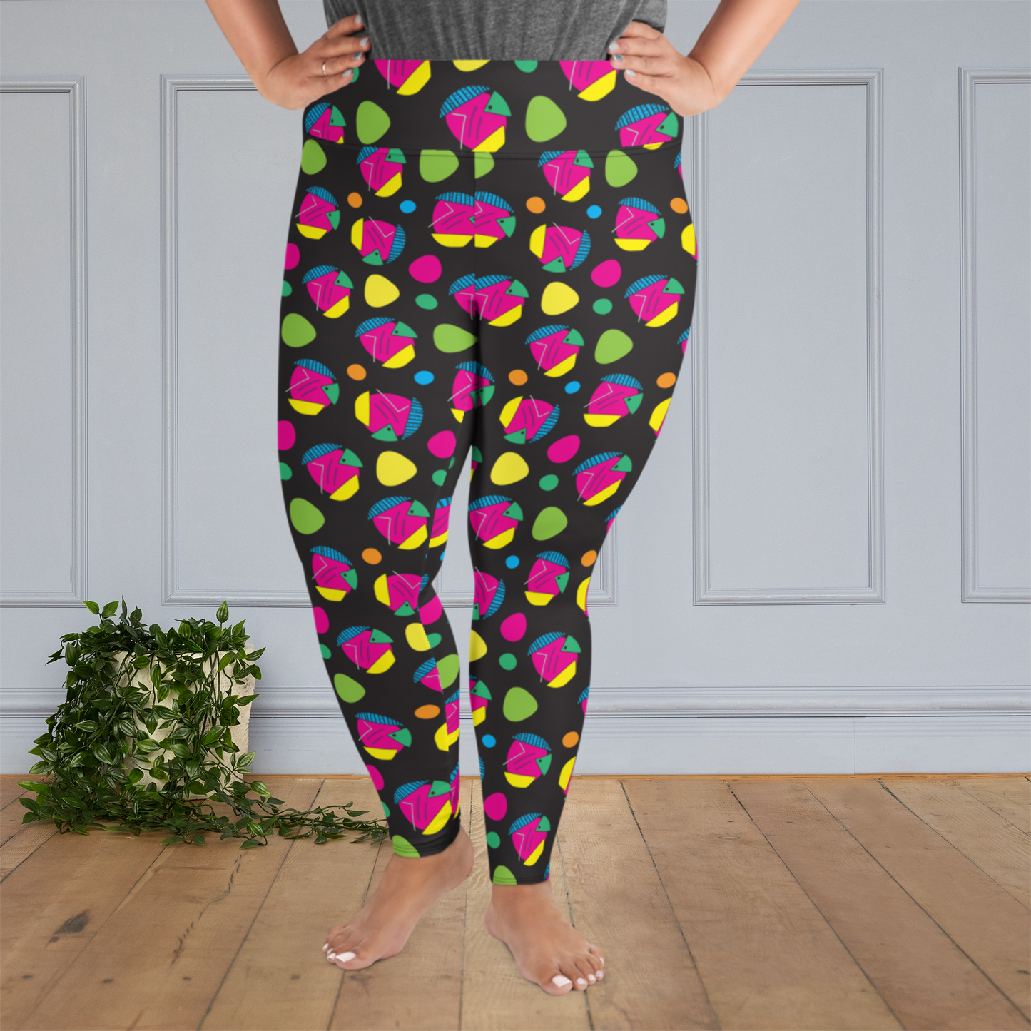 A curvy woman from the waist down wearing plus size high-waist Funk Party funky festival yoga leggings standing facing forwards with her hands on her hips. The woman is standing on a wooden floor with a white panelled wall in the background and a trailing plant on the floor. The funk party pattern  is an abstract pattern in vibrant pink, orange, yellow, green and blue in a spotty type arrangement on a black background