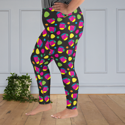 A curvy woman from the waist down wearing plus size high-waist Funk Party funky festival yoga leggings standing facing to the left with her feet slightly apart and with her hands on her hips. The woman is standing on a wooden floor with a white panelled wall in the background and a trailing plant on the floor. The funk party pattern  is an abstract pattern in vibrant pink, orange, yellow, green and blue in a spotty type arrangement on a black background