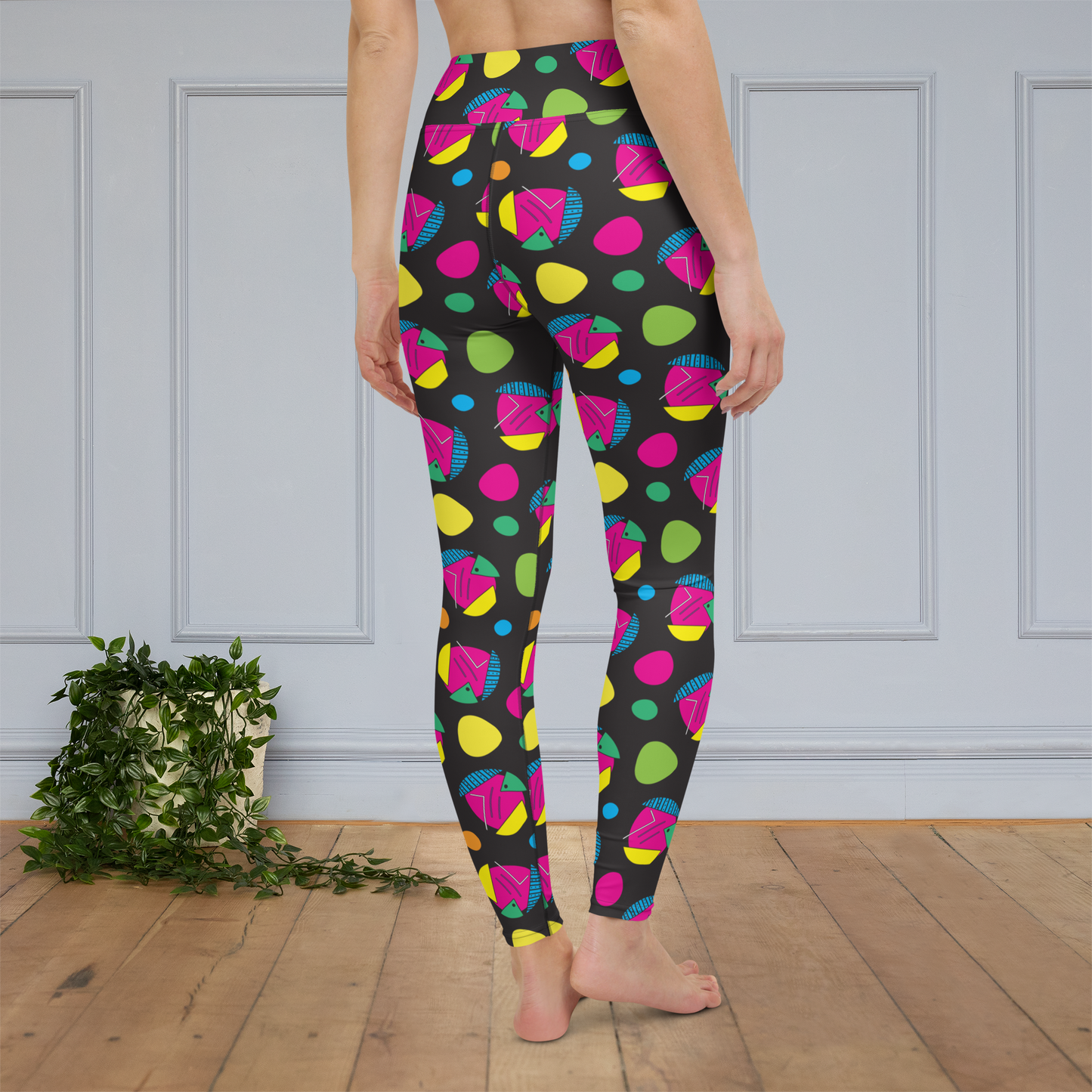 Woman from the waist down wearing high-waist Funk Party funky festival yoga leggings standing facing backwards. The woman is standing on a wooden floor with a white panelled wall in the background and a trailing plant on the floor. The funk party pattern  is an abstract pattern in vibrant pink, orange, yellow, green and blue in a spotty type arrangement on a black background