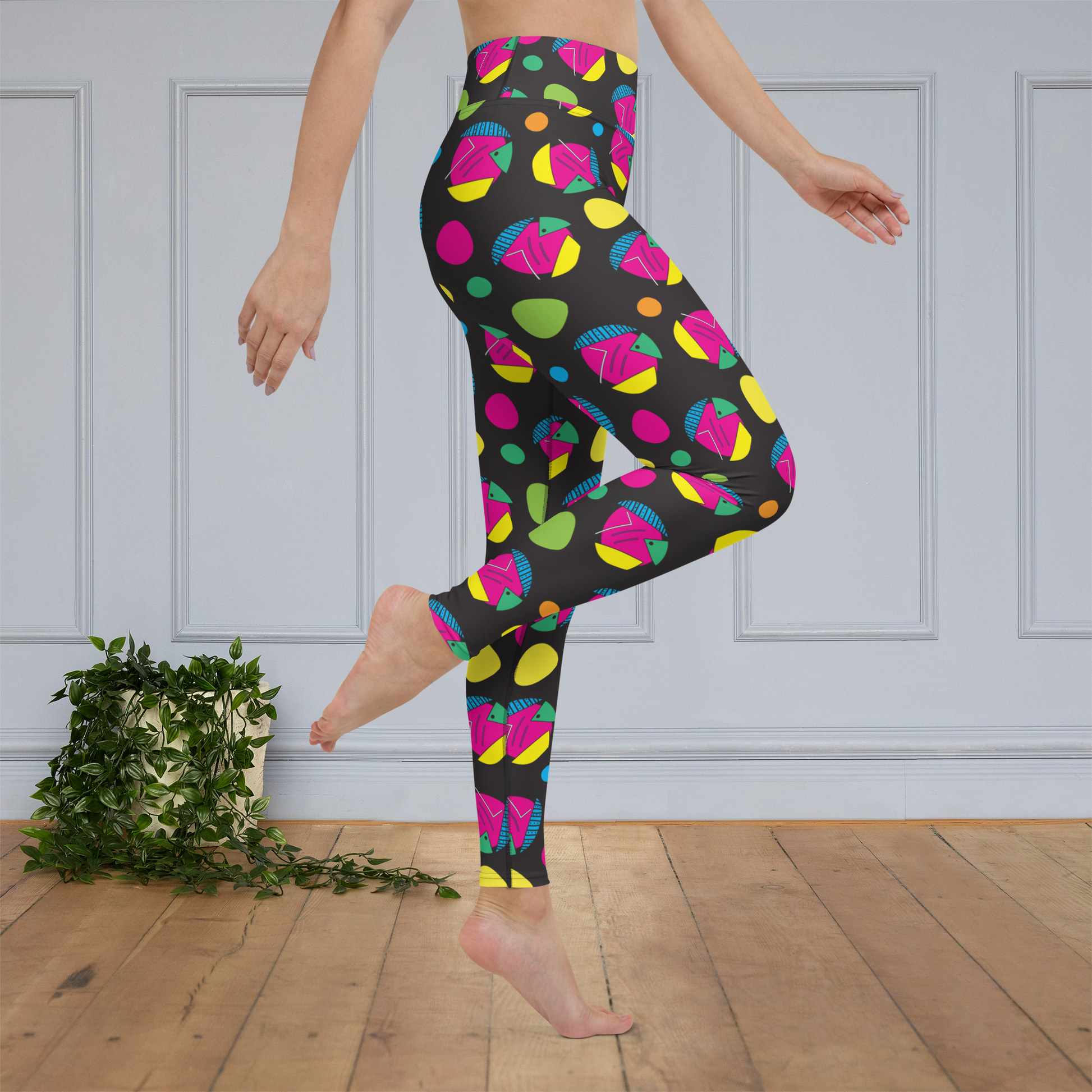 Woman from the waist down wearing high-waist Funk Party funky festival yoga leggings standing sideways on her left leg and right leg raised and bent with pointed toe. The woman is standing on a wooden floor with a white panelled wall in the background and a trailing plant on the floor. The funk party pattern  is an abstract pattern in vibrant pink, orange, yellow, green and blue in a spotty type arrangement on a black background