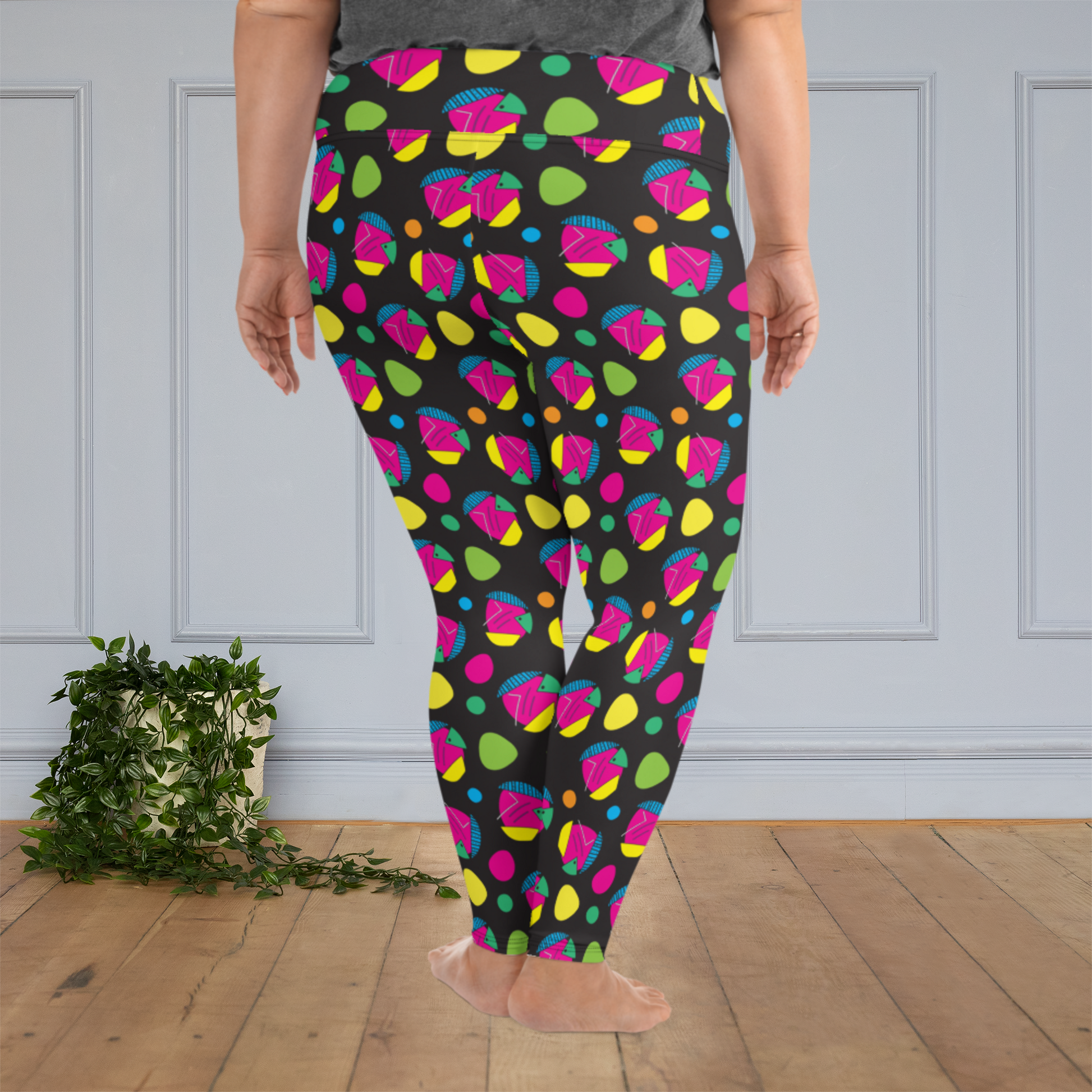A curvy woman from the waist down wearing plus size high-waist Funk Party funky festival yoga leggings standing facing to the back with her hands by her side. The woman is standing on a wooden floor with a white panelled wall in the background and a trailing plant on the floor. The funk party pattern  is an abstract pattern in vibrant pink, orange, yellow, green and blue in a spotty type arrangement on a black background