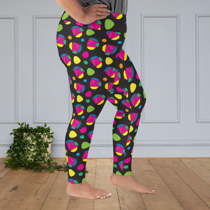 A curvy woman from the waist down wearing plus size high-waist Funk Party funky festival yoga leggings standing facing to the  right with her feet slightly apart and with her hands on her hips. The woman is standing on a wooden floor with a white panelled wall in the background and a trailing plant on the floor. The funk party pattern  is an abstract pattern in vibrant pink, orange, yellow, green and blue in a spotty type arrangement on a black background