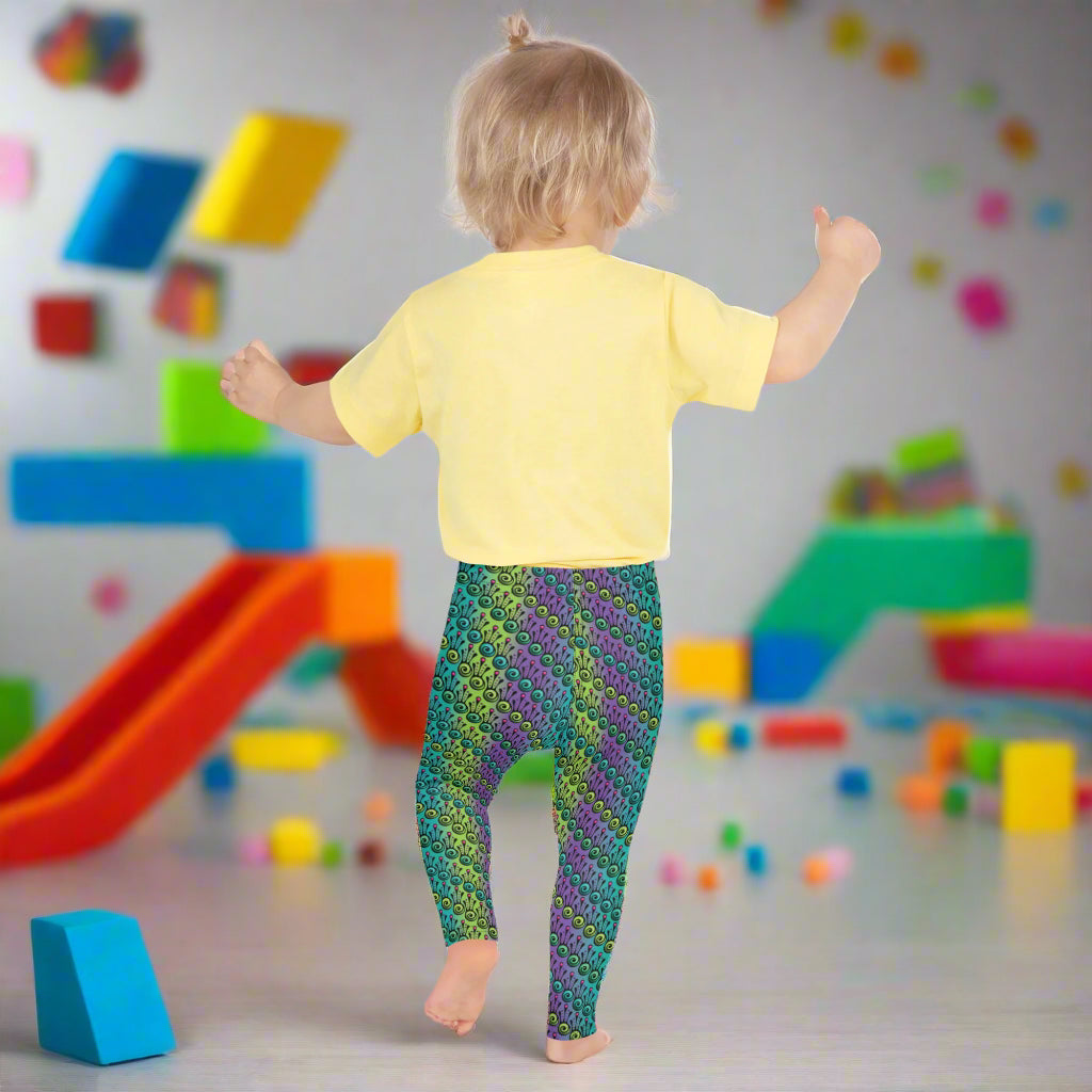 A toddler wearing kids size jungle tribal frog funky festival leggings and a pale yellow t-shirt stands facing away. They are in a play room filled with coloured blocks. The jungle tribal frog pattern is an abstract pattern with a blended green and purple background.