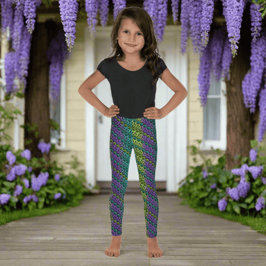 A young girl wearing kids size jungle tribal frog funky festival leggings, stands facing forwards with her hands on her hips. She is smiling and standing in front of a white door to a country house, with blooming wisteria flowers draping over the house. The jungle tribal frog pattern  is an abstract pattern with a blended green and purple background.