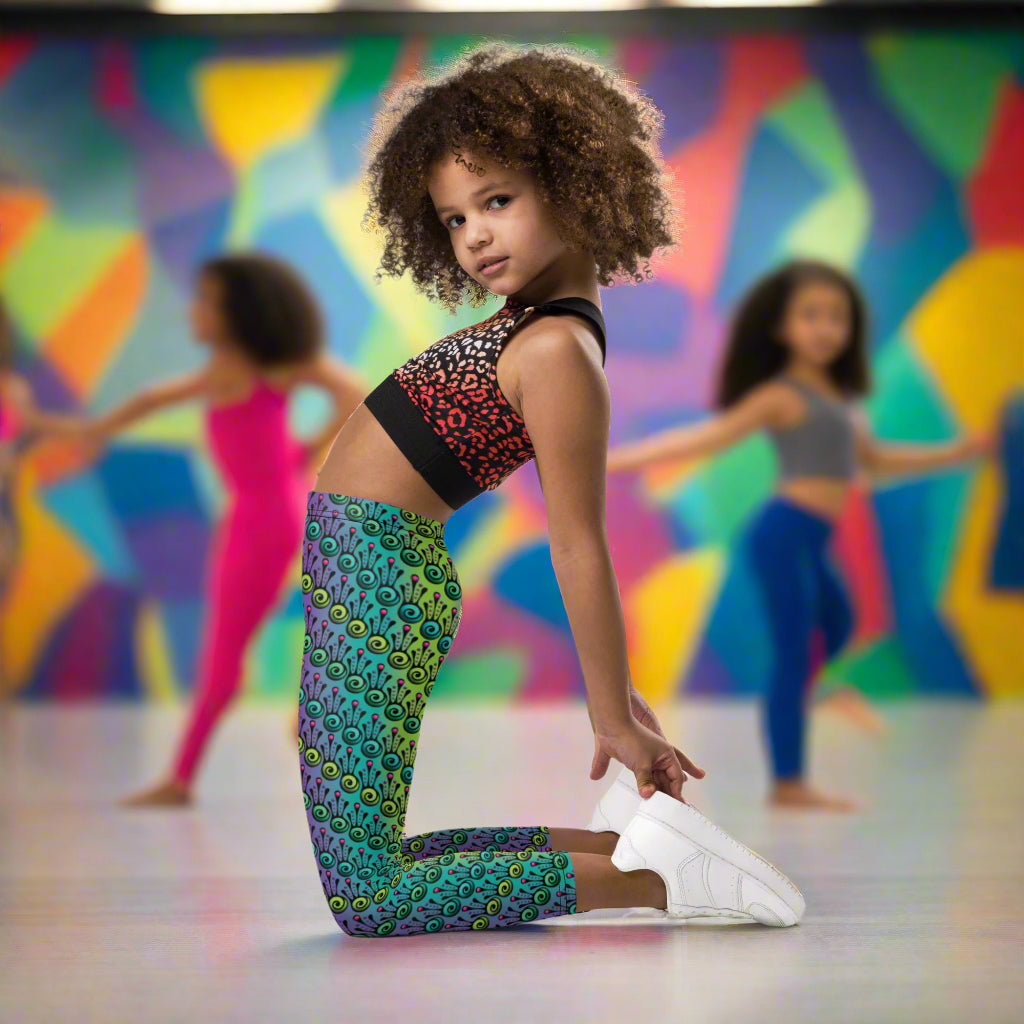 A young girl  poses wearing kids size jungle tribal frog funky festival leggings. She is in a brightly coloured dance studio with other young dancers in the background. The back wall is painted in vibrant multicoloured abstract shapes. The jungle tribal frog pattern  is an abstract pattern with a blended green and purple background.