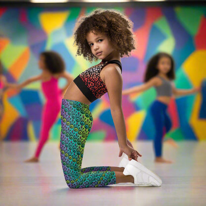 A young girl  poses wearing kids size jungle tribal frog funky festival leggings. She is in a brightly coloured dance studio with other young dancers in the background. The back wall is painted in vibrant multicoloured abstract shapes. The jungle tribal frog pattern  is an abstract pattern with a blended green and purple background.