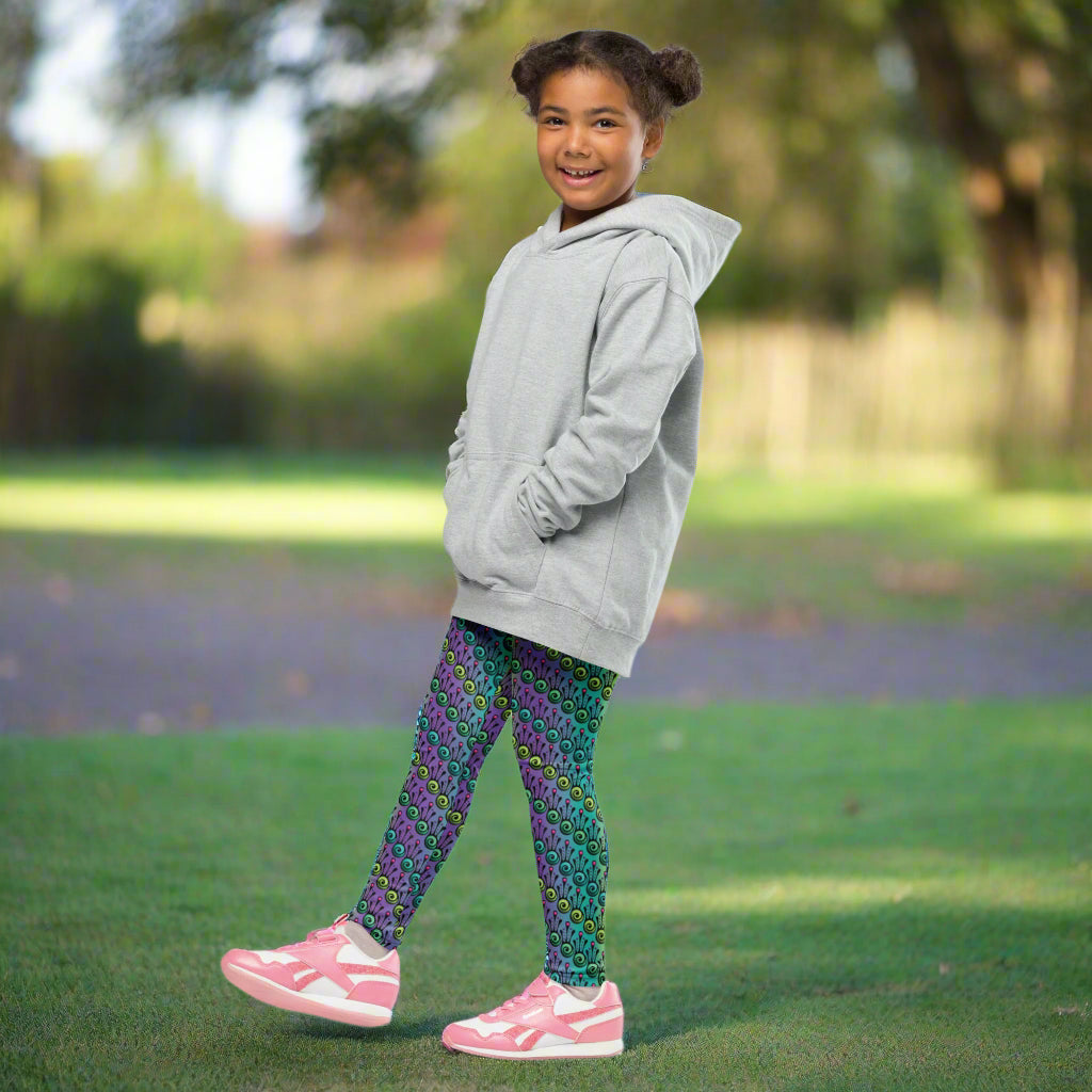 A young girl wearing kids size jungle tribal frog funky festival leggings and a grey oversized hoodie, stands facing the left with her right leg forward. She is in a park on a sunny day with trees in the background. The jungle tribal frog pattern  is an abstract pattern with a blended green and purple background.