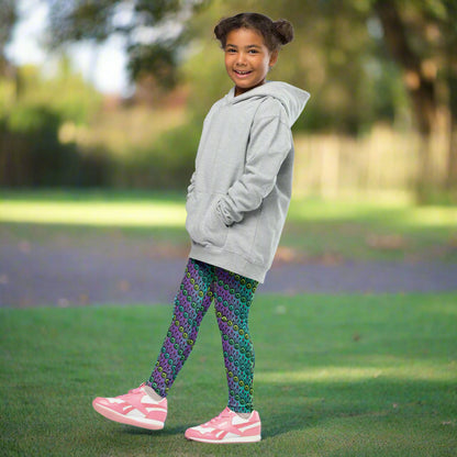 A young girl wearing kids size jungle tribal frog funky festival leggings and a grey oversized hoodie, stands facing the left with her right leg forward. She is in a park on a sunny day with trees in the background. The jungle tribal frog pattern  is an abstract pattern with a blended green and purple background.