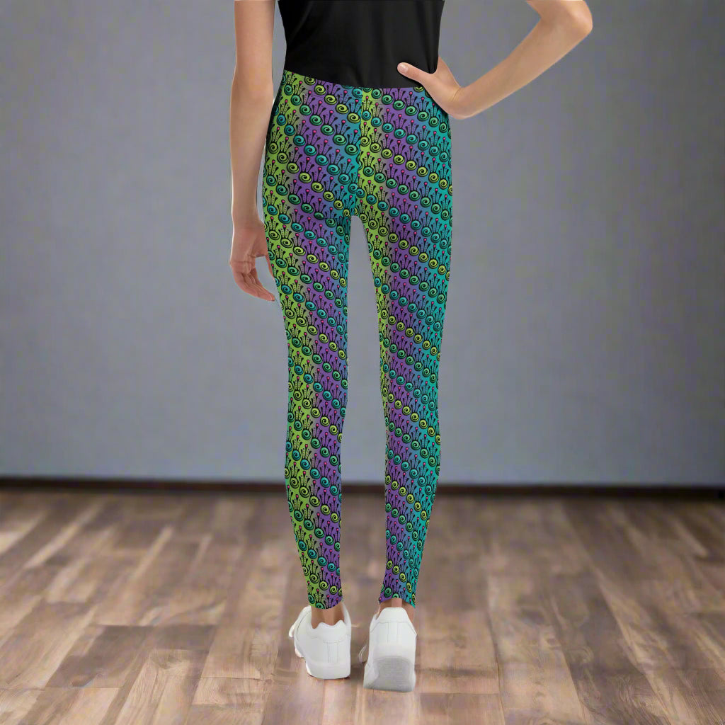 A youth pictured from the waist down wearing youth size jungle tribal frog funky festival leggings standing facing away with their right hand on their hip. They are wearing white trainers and standing on a wooden dance studio floor against a light grey wall. The jungle tribal frog pattern  is an abstract pattern with a blended green and purple background.