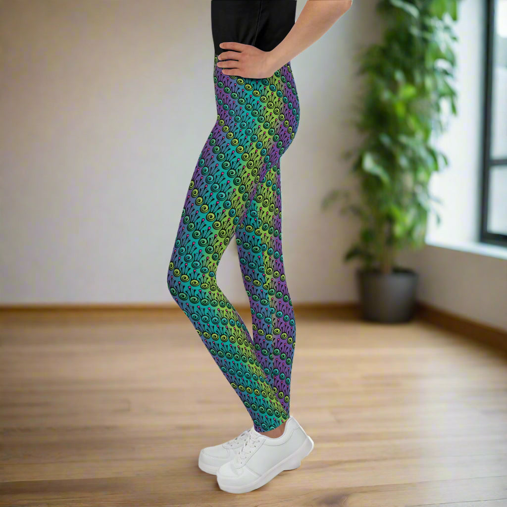 A youth pictured from the waist down wearing youth size jungle tribal frog funky festival leggings standing facing left with their hands on their hips. They are wearing white trainers and standing on a wooden dance studio floor with light streaming in from a large window on the right. A tall leafy plant sits by the window.  The jungle tribal frog pattern  is an abstract pattern with a blended green and purple background.
