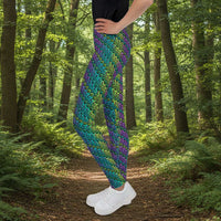 A youth pictured from the waist down wearing youth size jungle tribal frog funky festival leggings standing facing left with their hands on their hips. They are wearing white trainers and standing on a wooden dance studio floor with light streaming in from a large window on the right. A tall leafy plant sits by the window.  The jungle tribal frog pattern  is an abstract pattern with a blended green and purple background.