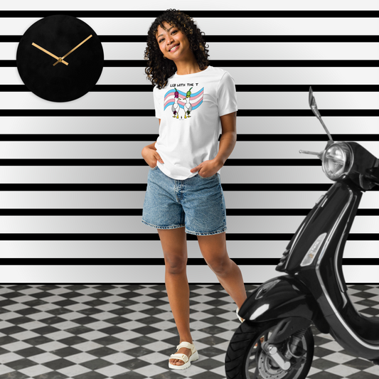 Woman posing casually facing forwards, wearing a fitted white Sassy Seagull LGB with the T LGBTQ+ Pride T-Shirt and  stonewash blue jean shorts. She is standing in a black and white theme space with horizontal stripes on the wall and a black moped to her right. The t-shirt is white with short sleeves. The graphic shows two cartoon style seagulls wearing colourful hats walking away in front of an trans flag, underneath the wording LGB with the T. It is a soft playful design for everyday wear