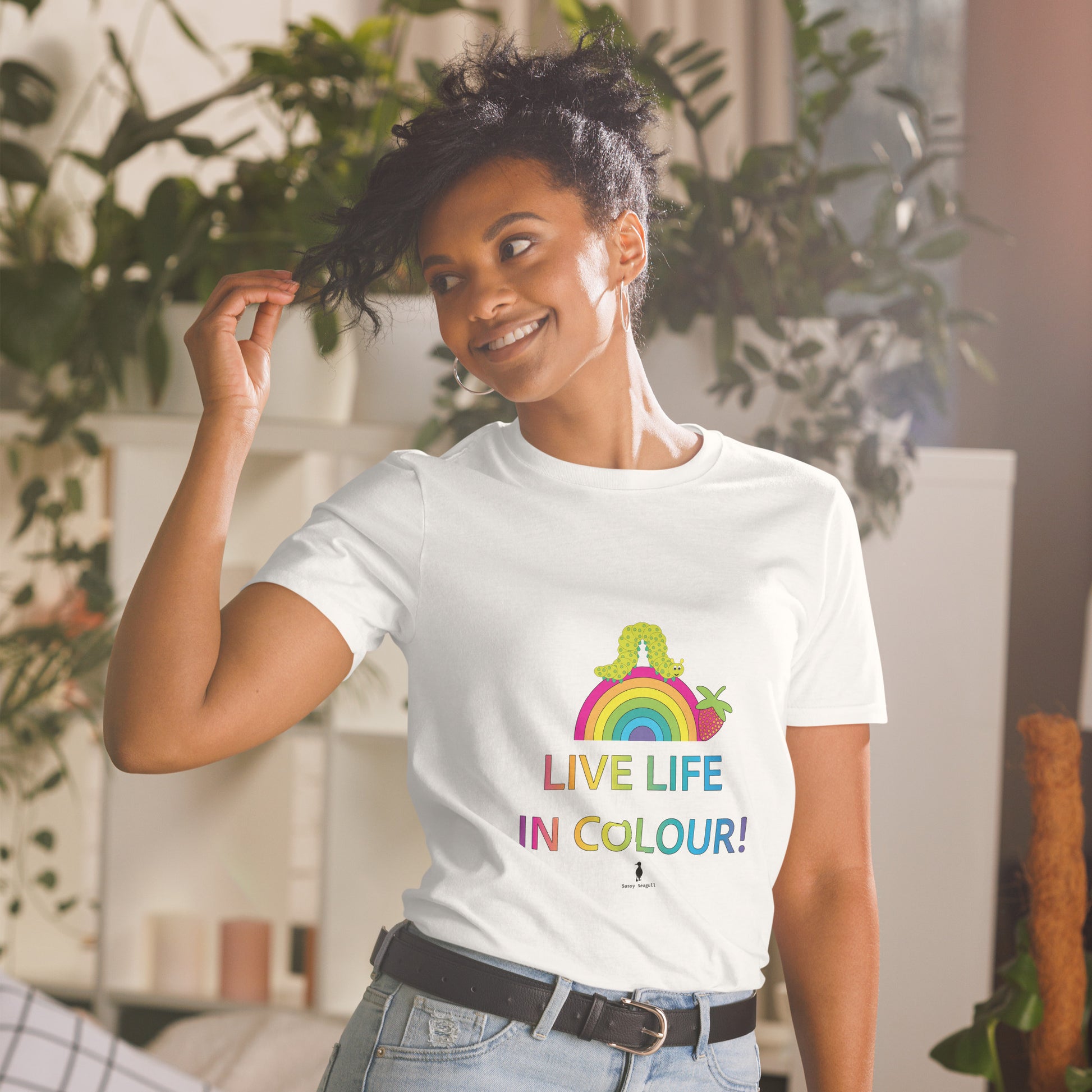 Woman wearing a white 100% cotton vibrant, rainbow live life in colour t-shirt in a bohemian interior decorated with plants.