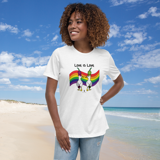 Woman standing facing forwards and looking to the left, wearing a fitted white Sassy Seagull Love Is Love LGBTQ+ Pride T-Shirt and pale stonewash blue jeans. She is standing on a white sandy beach with the sea in the distance. There are white clouds in a blue sky. The t-shirt is white with short sleeves. The graphic shows two cartoon style seagulls wearing colourful hats walking away in front of an LGBTQIA pride flag, underneath the wording Love Is Love. It is a bold playful design for everyday wear.