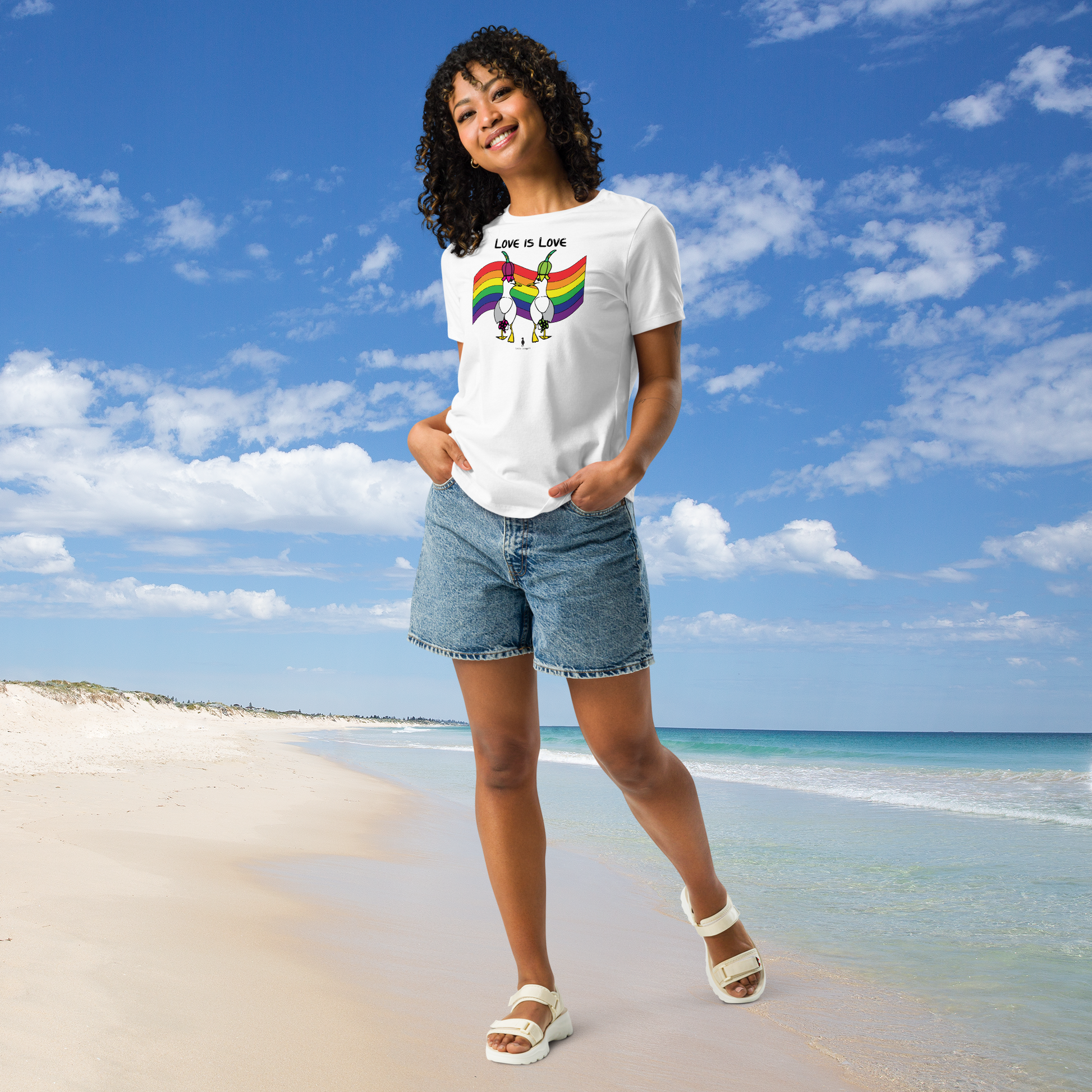Woman posing casually facing forwards, wearing a fitted white Sassy Seagull Love Is Love LGBTQ+ Pride T-Shirt and  stonewash blue jean shorts. She is standing on a white sandy beach with the sea in the distance. There are white clouds in a blue sky. The t-shirt is white with short sleeves. The graphic shows two cartoon style seagulls wearing colourful hats walking away in front of an LGBTQIA pride flag, underneath the wording Love Is Love. It is a bold playful design for everyday wear.
