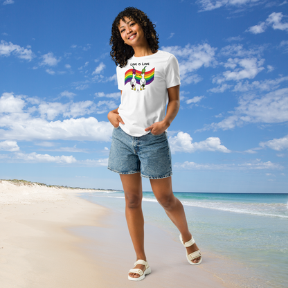 Woman posing casually facing forwards, wearing a fitted white Sassy Seagull Love Is Love LGBTQ+ Pride T-Shirt and  stonewash blue jean shorts. She is standing on a white sandy beach with the sea in the distance. There are white clouds in a blue sky. The t-shirt is white with short sleeves. The graphic shows two cartoon style seagulls wearing colourful hats walking away in front of an LGBTQIA pride flag, underneath the wording Love Is Love. It is a bold playful design for everyday wear.