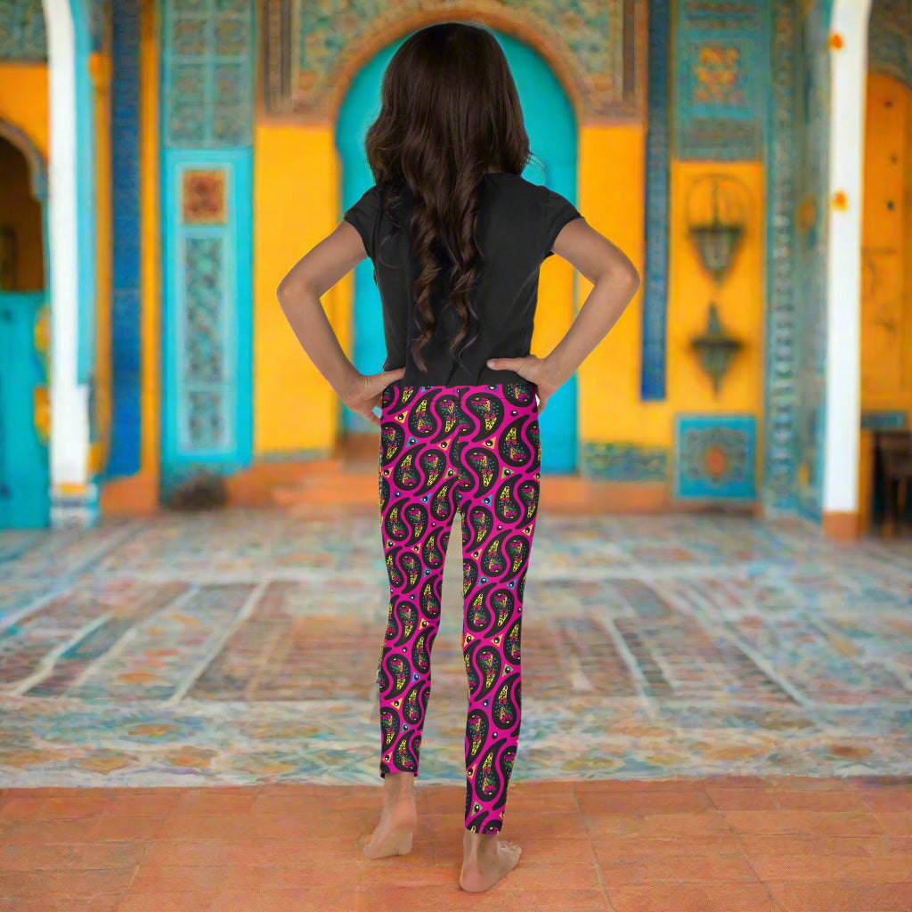 A young girl poses facing to the back inside an ornate orange, yellow and turquoise moroccan-style building with ornate  tiled floor. She is wearing pink paisley party funky festival leggings, and a black t-shirt. The paisley party pattern  is a colourful repeating large paisley pattern, made up of vibrant colours including pink, orange, yellow, green, blue and purple. In between the paisley print are colourful triangles in the same colours. The pattern  background is hot pink
