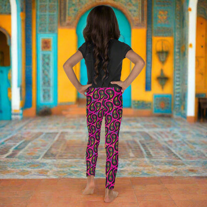 A young girl poses facing to the back inside an ornate orange, yellow and turquoise moroccan-style building with ornate  tiled floor. She is wearing pink paisley party funky festival leggings, and a black t-shirt. The paisley party pattern  is a colourful repeating large paisley pattern, made up of vibrant colours including pink, orange, yellow, green, blue and purple. In between the paisley print are colourful triangles in the same colours. The pattern  background is hot pink