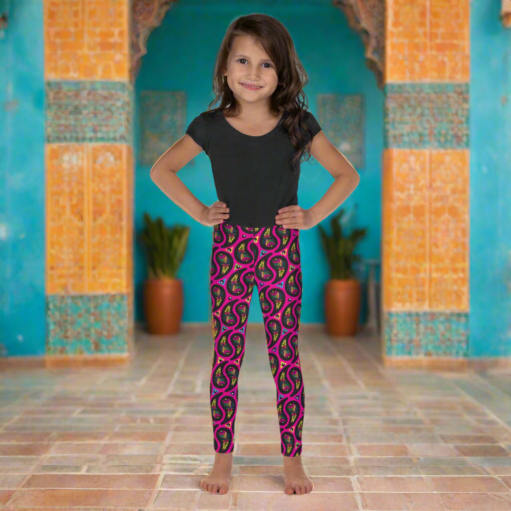 A young girl poses facing to the front outside an ornate orange and turquoise moroccan-style building with an arch and tiled floor. She is wearing pink paisley party funky festival leggings, and a black t-shirt. The paisley party pattern  is a colourful repeating large paisley pattern, made up of vibrant colours including pink, orange, yellow, green, blue and purple. In between the paisley print are colourful triangles in the same colours. The pattern  background is hot pink