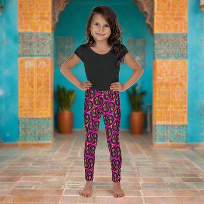 A young girl poses facing to the front outside an ornate orange and turquoise moroccan-style building with an arch and tiled floor. She is wearing pink paisley party funky festival leggings, and a black t-shirt. The paisley party pattern  is a colourful repeating large paisley pattern, made up of vibrant colours including pink, orange, yellow, green, blue and purple. In between the paisley print are colourful triangles in the same colours. The pattern  background is hot pink