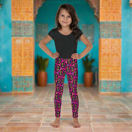 A young girl poses facing to the front outside an ornate orange and turquoise moroccan-style building with an arch and tiled floor. She is wearing pink paisley party funky festival leggings, and a black t-shirt. The paisley party pattern  is a colourful repeating large paisley pattern, made up of vibrant colours including pink, orange, yellow, green, blue and purple. In between the paisley print are colourful triangles in the same colours. The pattern  background is hot pink