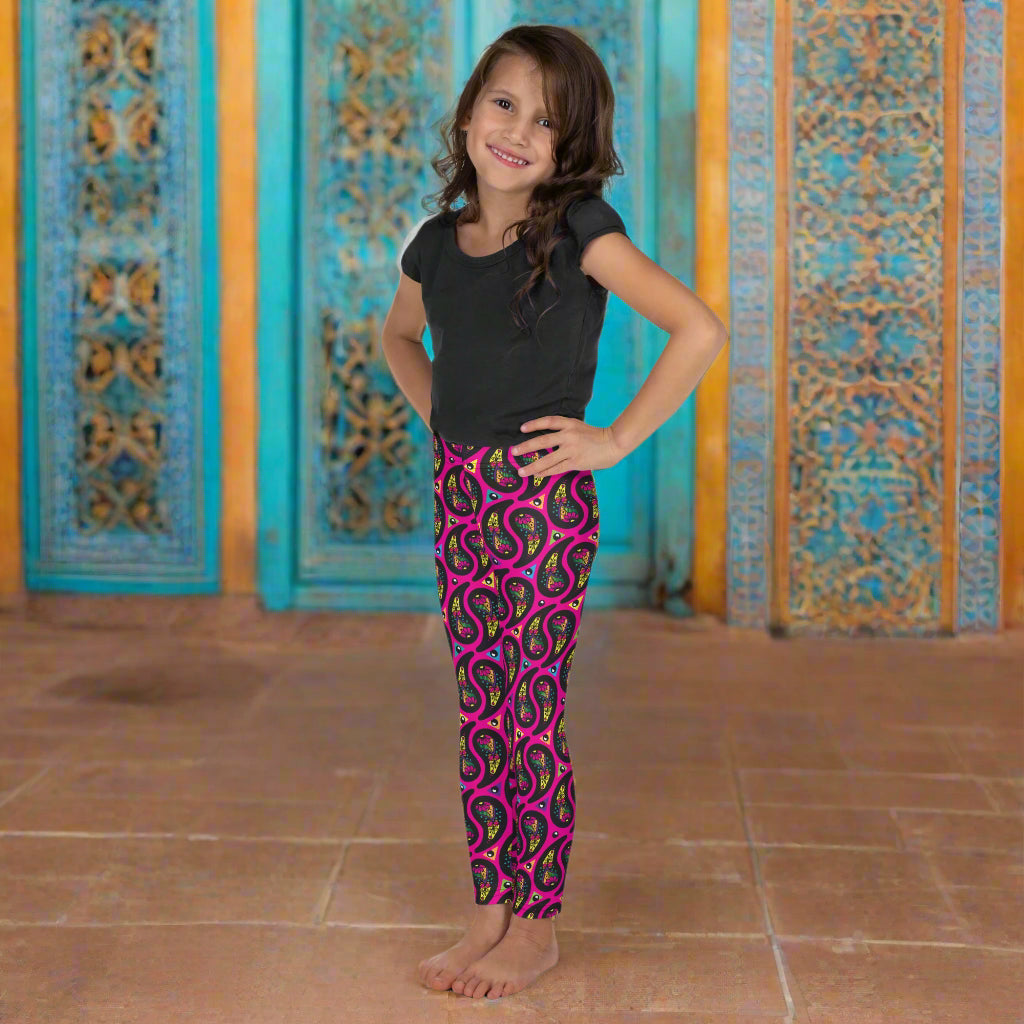 A young girl poses facing to the left inside an ornate orange and turquoise moroccan-style building with a tiled floor. She is wearing pink paisley party funky festival leggings, and a black t-shirt. The paisley party pattern  is a colourful repeating large paisley pattern, made up of vibrant colours including pink, orange, yellow, green, blue and purple. In between the paisley print are colourful triangles in the same colours. The pattern  background is hot pink