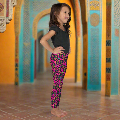 A young girl poses facing to the right inside an ornate orange and turquoise moroccan-style building with arches and a tiled floor. She is wearing pink paisley party funky festival leggings, and a black t-shirt. The paisley party pattern  is a colourful repeating large paisley pattern, made up of vibrant colours including pink, orange, yellow, green, blue and purple. In between the paisley print are colourful triangles in the same colours. The pattern  background is hot pink