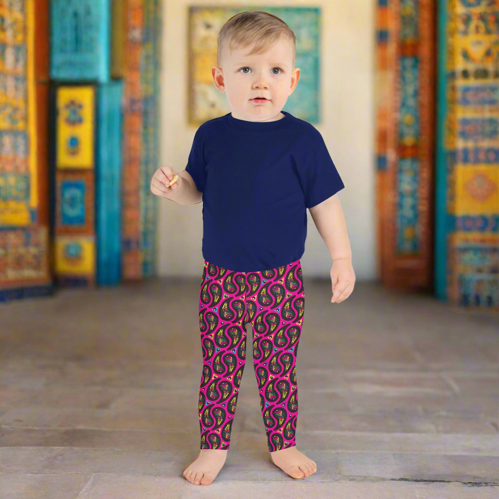A toddler poses facing forward inside an ornate orange and turquoise moroccan-style building with a tiled floor. He is wearing pink paisley party funky festival leggings, and a navy t-shirt. The paisley party pattern is a colourful repeating large paisley pattern, made up of vibrant colours including pink, orange, yellow, green, blue and purple. In between the paisley print are colourful triangles in the same colours. The pattern  background is hot pink