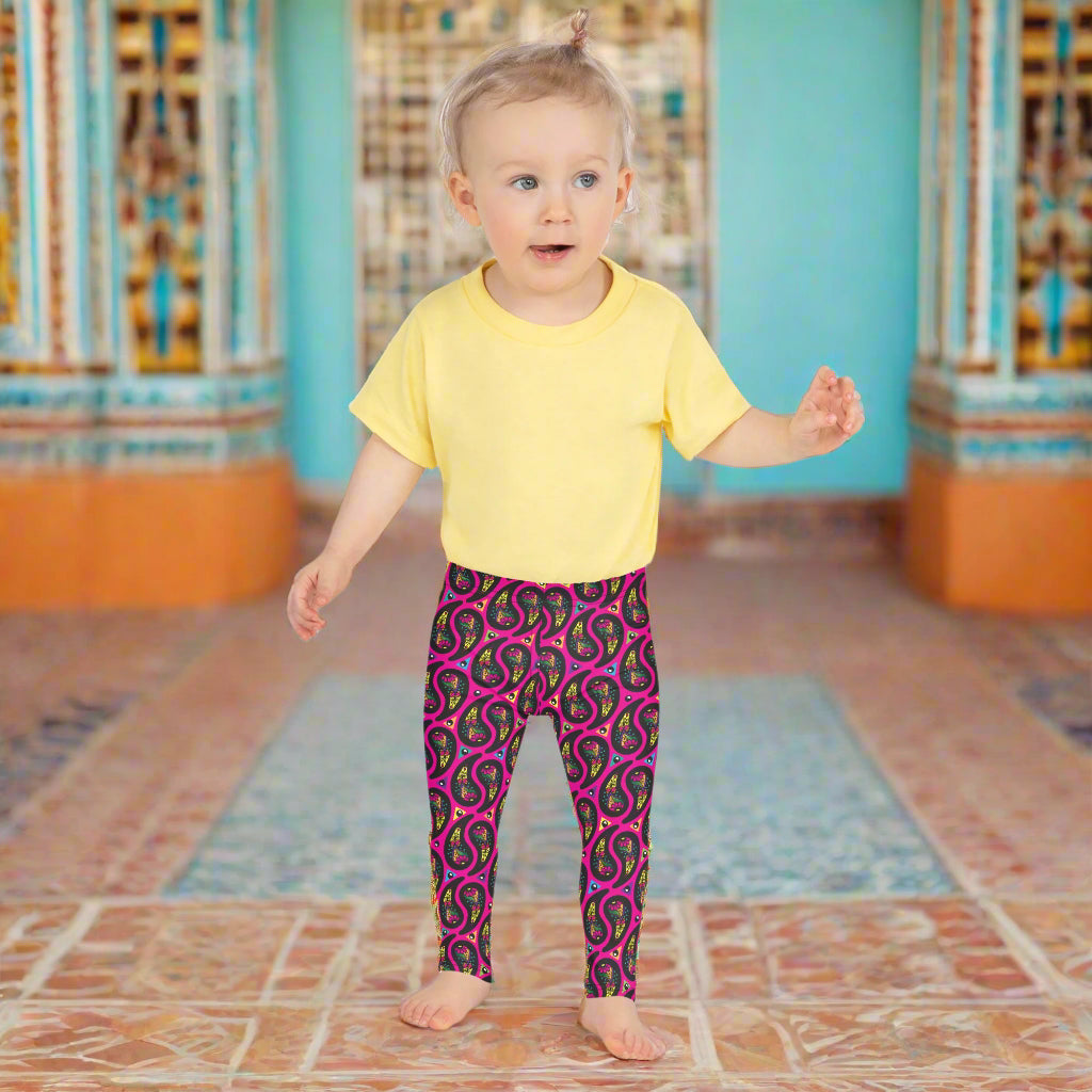 A toddler poses facing forward inside a decorative orange and turquoise moroccan-style building with a tiled floor. She is wearing pink paisley party funky festival leggings, and a pale yellow t-shirt. The paisley party pattern is a colourful repeating large paisley pattern, made up of vibrant colours including pink, orange, yellow, green, blue and purple. In between the paisley print are colourful triangles in the same colours. The pattern  background is hot pink