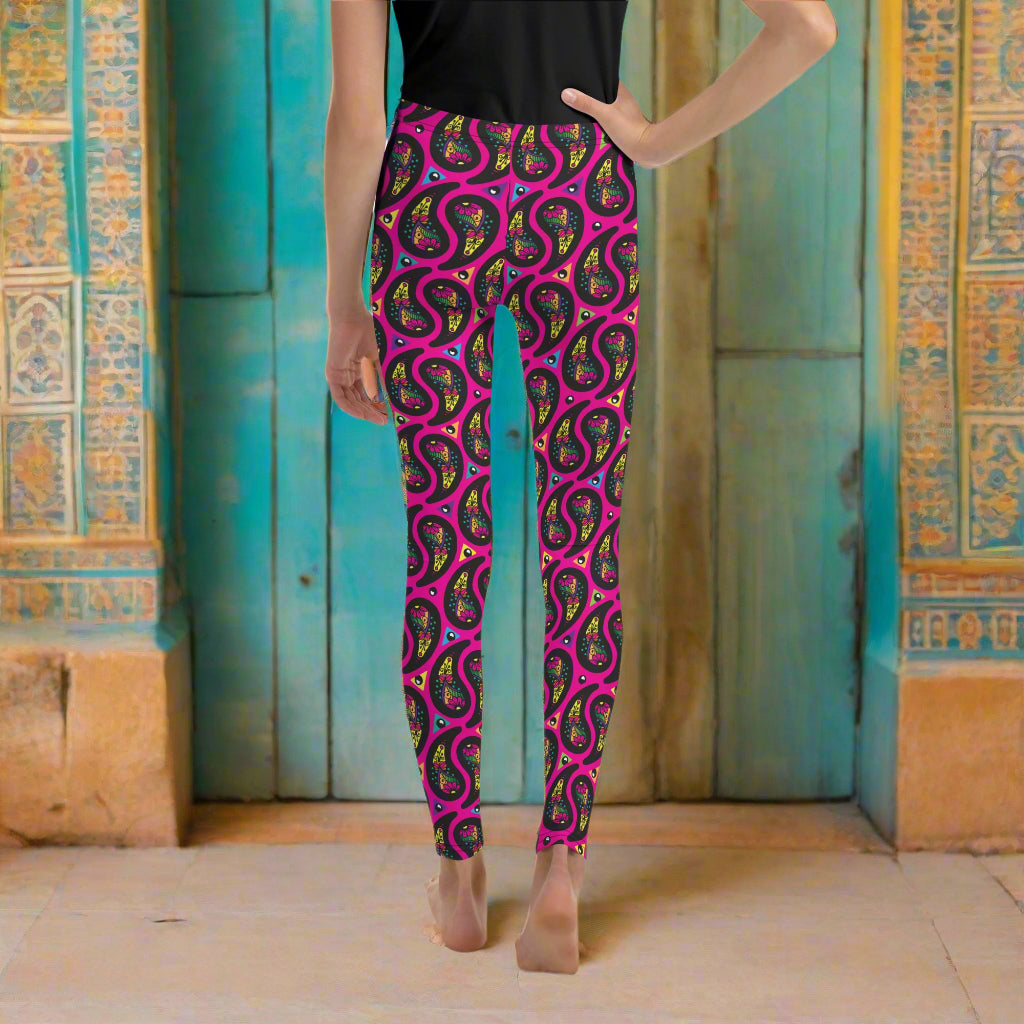 A young person from the waist down poses facing away towards an decorative orange and turquoise moroccan-style building. They are wearing pink paisley party funky festival leggings. The paisley party pattern is a colourful repeating large paisley pattern, made up of vibrant colours including pink, orange, yellow, green, blue and purple. In between the paisley print are colourful triangles in the same colours. The pattern  background is hot pink