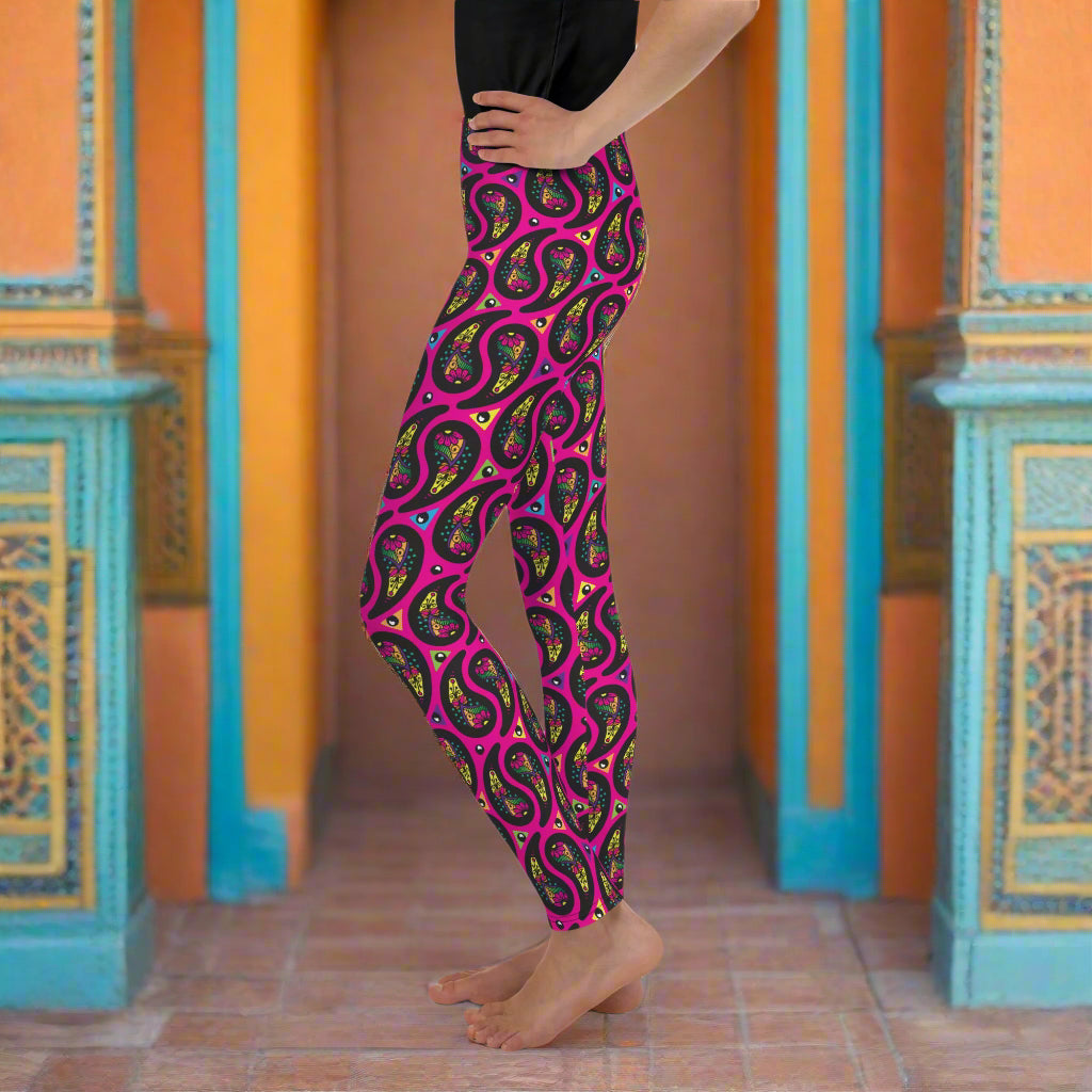 A young person from the waist down poses facing to the left inside an ornate orange and turquoise moroccan-style building with a tiled floor. They are wearing pink paisley party funky festival leggings. The paisley party pattern is a colourful repeating large paisley pattern, made up of vibrant colours including pink, orange, yellow, green, blue and purple. In between the paisley print are colourful triangles in the same colours. The pattern  background is hot pink