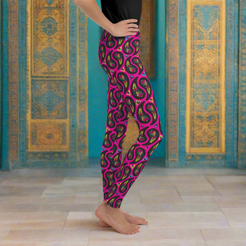 A young person from the waist down poses facing to the right inside an ornate gold and turquoise moroccan-style building with a tiled floor. They are wearing pink paisley party funky festival leggings. The paisley party pattern is a colourful repeating large paisley pattern, made up of vibrant colours including pink, orange, yellow, green, blue and purple. In between the paisley print are colourful triangles in the same colours. The pattern  background is hot pink