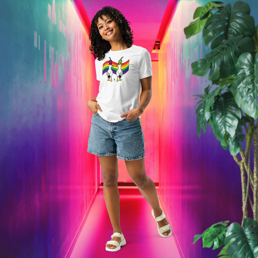 Woman posing casually facing forwards, wearing a fitted white Sassy Seagull Pride Flag LGBTQ+ Pride T-Shirt and stonewash blue jean shorts. She is standing in a rainbow coloured corridor in pinks, purples, blues and oranges with a tall green leafy plant to the front right. The t-shirt is white with short sleeves. The graphic shows two cartoon style seagulls wearing colourful hats walking away in front of an LGBTQIA pride flag. It is a bold playful design for everyday wear