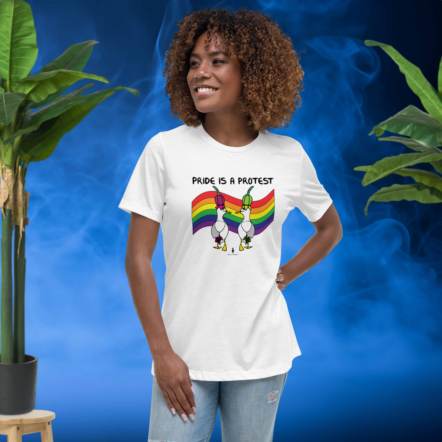 Woman standing facing forwards and looking to the left, wearing a fitted white Sassy Seagull Pride is a Protest LGBTQ+ Pride T-Shirt and pale stonewash blue jeans. She is standing in front of a smoky blue background with plants either side. The t-shirt is white with short sleeves. The graphic shows two cartoon style seagulls wearing colourful hats walking away in front of an LGBTQIA pride flag, underneath the wording Pride is a Protest. It is a bold playful design for everyday wear.
