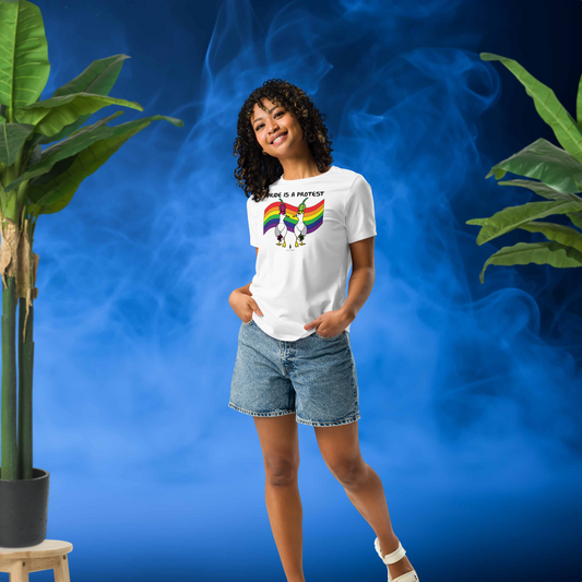 Woman posing casually facing forwards, wearing a fitted white Sassy Seagull Pride is a Protest LGBTQ+ Pride T-Shirt and  stonewash blue jean shorts. She is standing in front of a smoky blue background with plants either side. The t-shirt is white with short sleeves. The graphic shows two cartoon style seagulls wearing colourful hats walking away in front of an LGBTQIA pride flag, underneath the wording Pride is a Protest. It is a bold playful design for everyday wear
